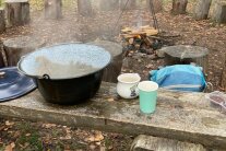 eine Holzbank mit Tassen und einem großen Topf voller Fichtennadeltee vor einem Lagerfeuer